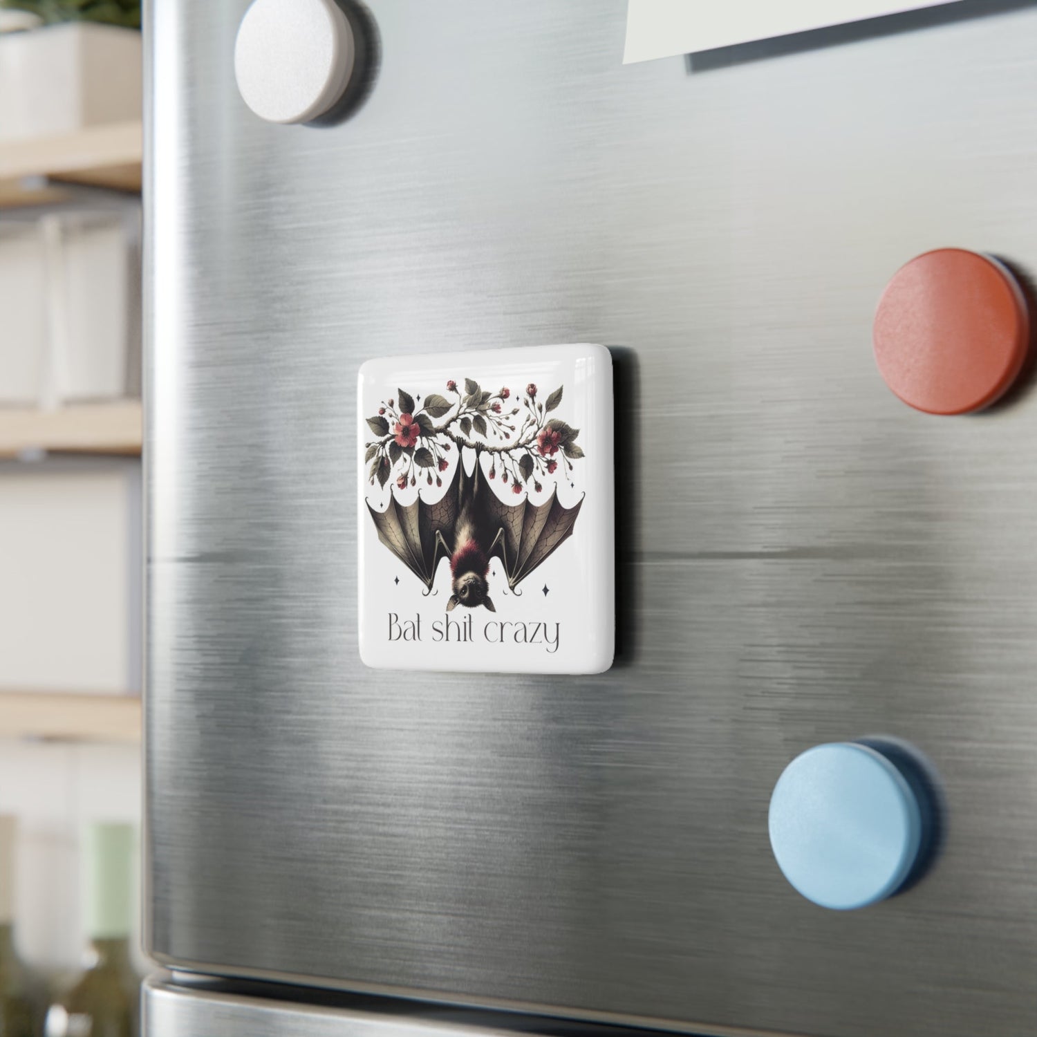 a square ceramic magnet on a refrigerator with a bat hanging upside down on a flower or rose bush that says bat shit crazy 